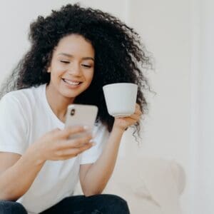 Peaceful curly woman scrolling social networks via modern mobile phone, watches funny videos